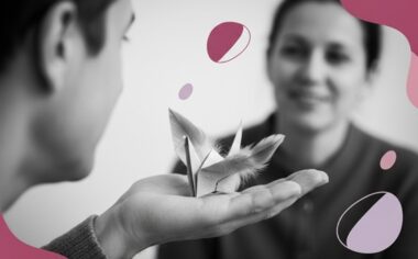 Un oiseau origami posé dans la paume d’une main ouverte, en noir et blanc avec touches rose et violet.