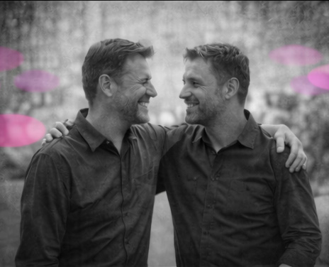 Deux hommes adultes se tenant par l’épaule, souriants, en noir et blanc, avec touches rose et violet. Exemple de liens