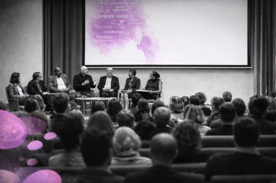 Panel de conférenciers lors d’un congrès, salle comble, image en noir et blanc avec touches de rose Mimethys.
