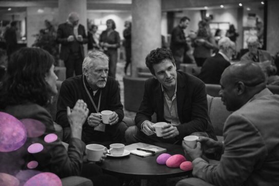 Participants en discussion lors d’un congrès de l’Institut Mimethys, en noir et blanc, avec des œufs roses et violets symboliques en premier plan.